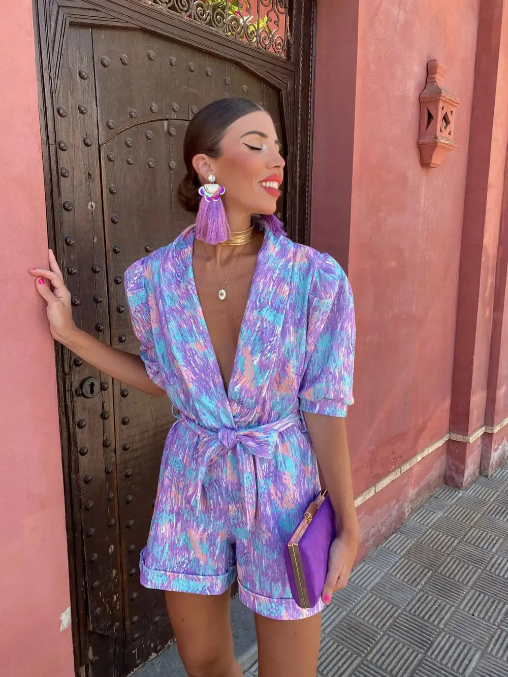 Women wearing a purple abstract metallic printed short sleeve romper with a tie waist, side view.