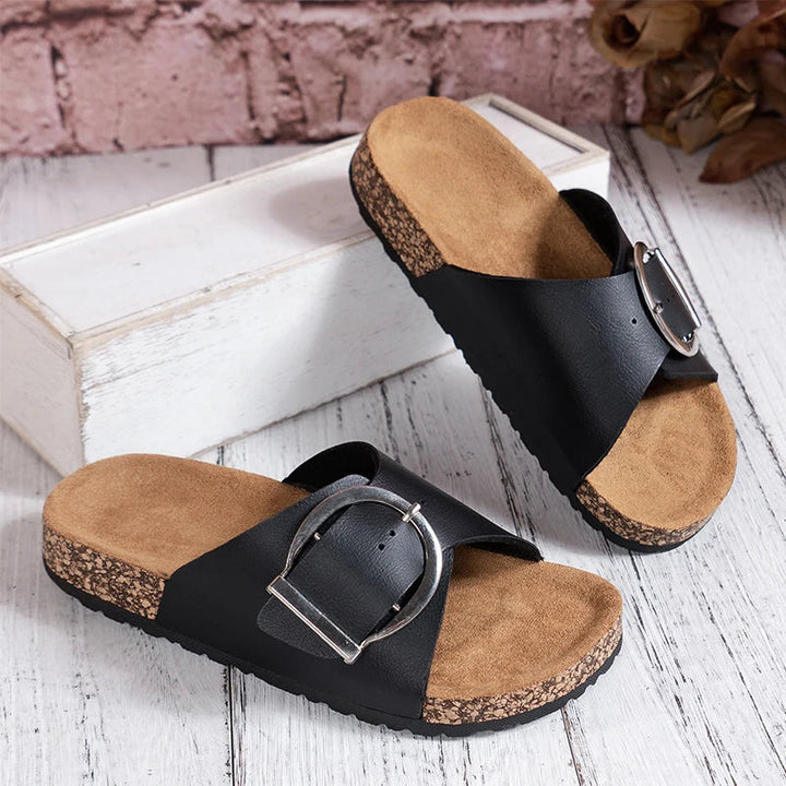 Pair of women black buckle strap slide mules with cork-look soles on a white wooden floor