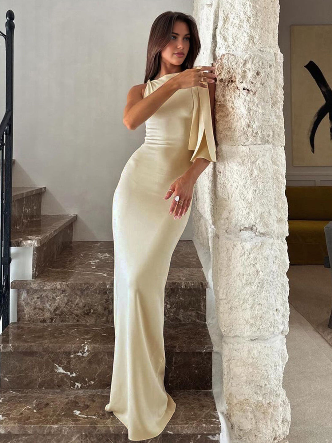 Women's off-white one-shoulder maxi dress, side profile, leaning against a pillar
