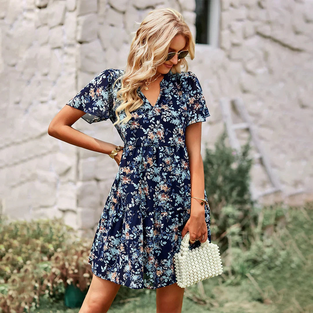 Women navy blue floral print mini sundress with V-neck and short flutter sleeves, standing by wall.