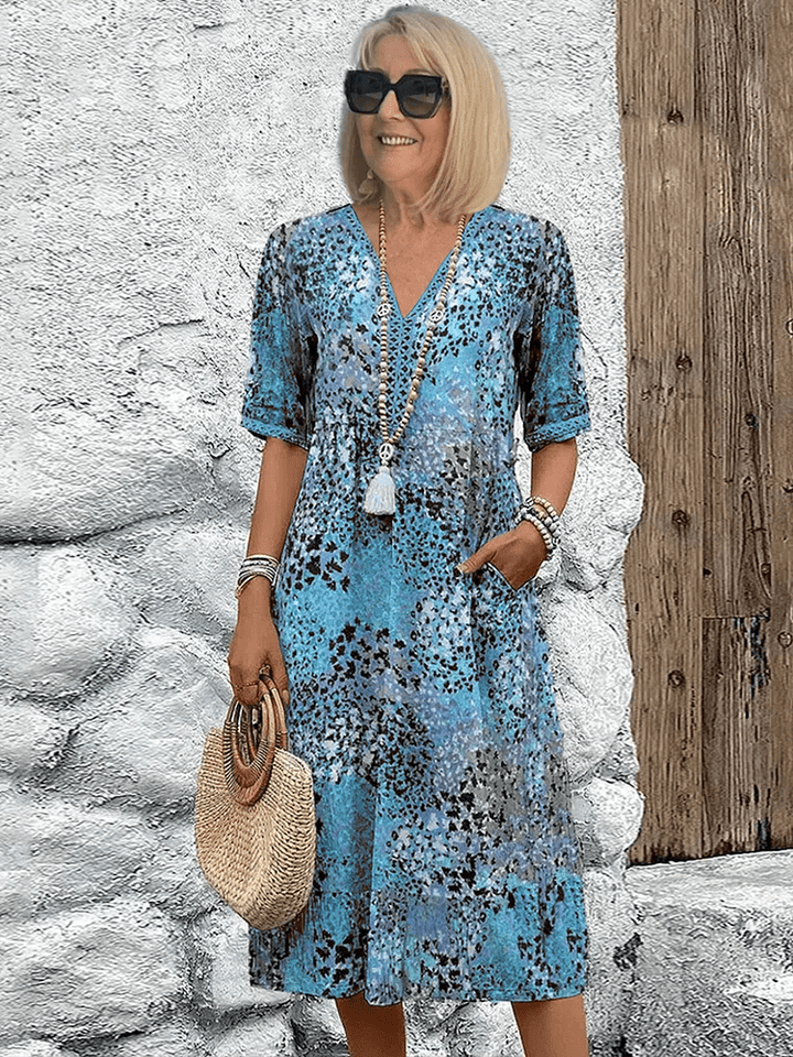 Women's blue multi-color printed midi dress with V-neckline and short sleeves, hands in pockets.