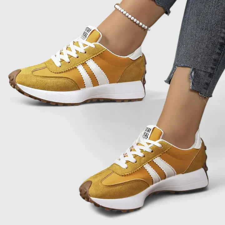 Women's yellow lace-up sneakers with white stripes, brown toe detail, worn with cuffed denim jeans